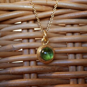Puede incluir: Un collar de tono dorado con una cadena fina y un colgante circular que presenta una piedra preciosa verde transl&uacute;cida engastada. El colgante muestra un acabado met&aacute;lico pulido y una piedra verde redondeada de color vibrante.