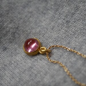 May include: A gold pendant necklace featuring a round, cabochon-cut pink gemstone set in a smooth gold bezel. The pendant is attached to a delicate gold chain with small, uniform links, showcasing a polished finish on the metal and stone.