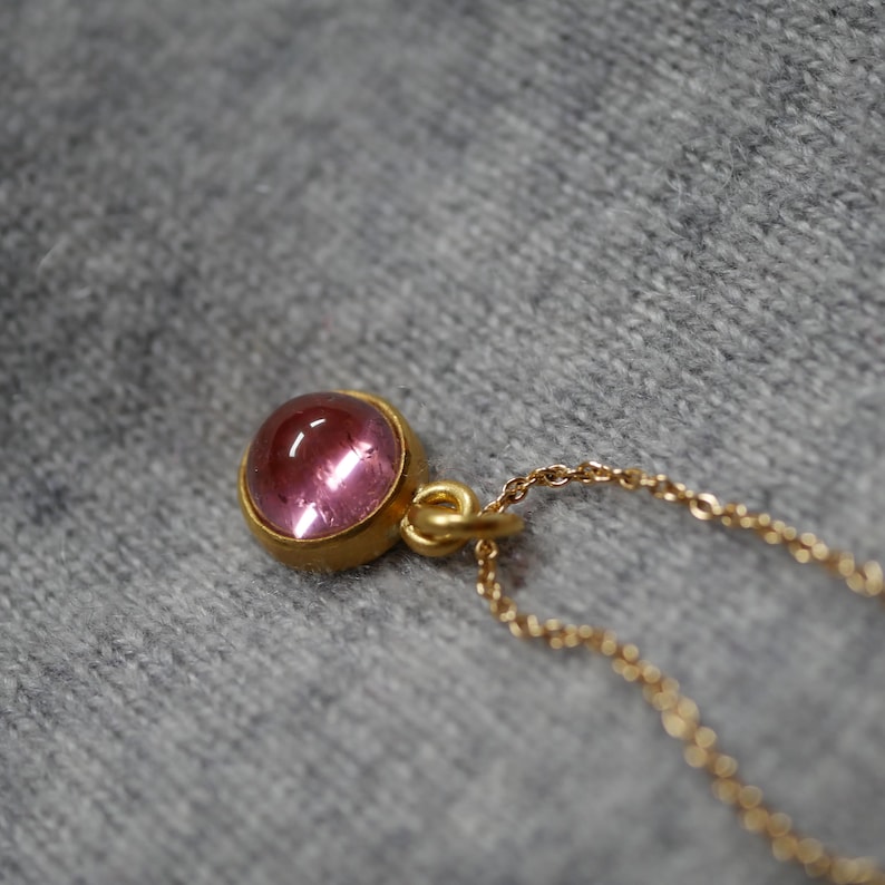 May include: A gold pendant necklace featuring a round, cabochon-cut pink gemstone set in a smooth gold bezel. The pendant is attached to a delicate gold chain with small, uniform links, showcasing a polished finish on the metal and stone.