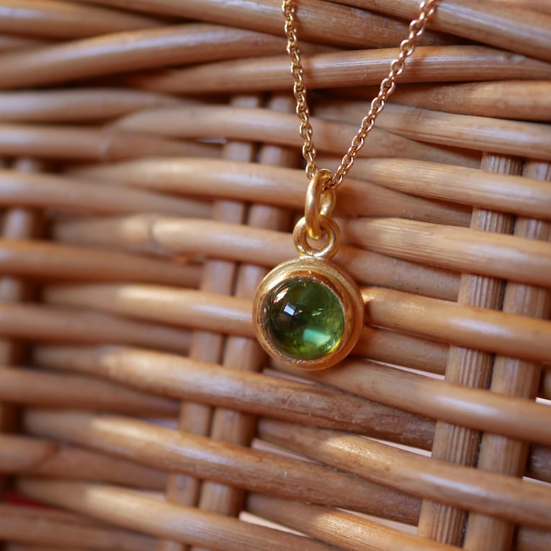 Puede incluir: Un collar de tono dorado con un peque&ntilde;o colgante redondo engastado con una piedra preciosa transl&uacute;cida de color verde oliva. El colgante cuelga de una delicada cadena de tono dorado, mostrando un dise&ntilde;o circular simple y pulido.