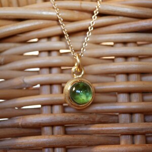 Puede incluir: Un collar de tono dorado con una cadena delicada y un colgante circular que presenta una piedra preciosa verde transl&uacute;cida, lisa y engastada en el centro. El colgante tiene un acabado met&aacute;lico pulido y cuelga de una cadena de eslabones fina.