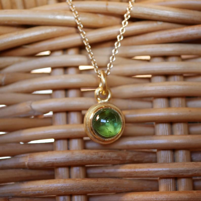 Puede incluir: Un collar de tono dorado con una cadena delicada y un colgante circular que presenta una piedra preciosa verde transl&uacute;cida, lisa y engastada en el centro. El colgante tiene un acabado met&aacute;lico pulido y cuelga de una cadena de eslabones fina.