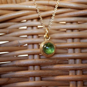 Puede incluir: Un collar de cadena dorada con un peque&ntilde;o colgante redondo de piedra preciosa verde en engaste de bisel. El colgante tiene un marco dorado liso y pulido que sujeta la piedra verde transl&uacute;cida y vibrante en el centro, suspendido de una delicada cadena de eslabones dorados.