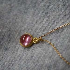 May include: A gold necklace featuring a small, round, cabochon-cut pink gemstone set in a simple gold bezel setting. The pendant is attached to a delicate gold chain, showcasing a smooth, polished surface and a vibrant, translucent pink hue.