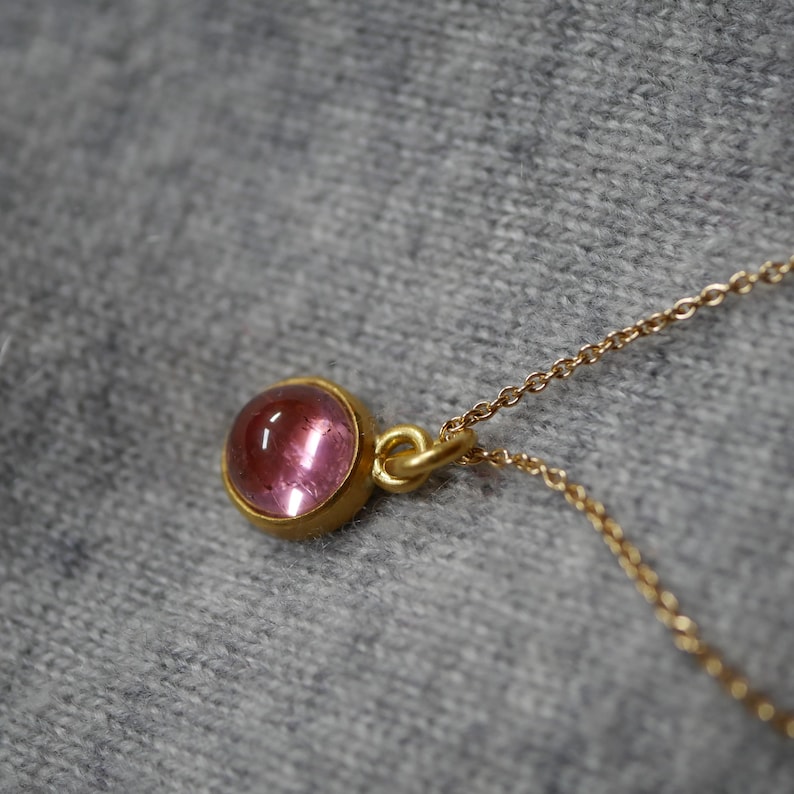 May include: A gold necklace featuring a small, round, cabochon-cut pink gemstone set in a simple gold bezel setting. The pendant is attached to a delicate gold chain, showcasing a smooth, polished surface and a vibrant, translucent pink hue.