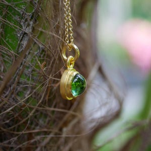 Puede incluir: Un collar con colgante de oro que presenta una peque&ntilde;a piedra preciosa verde de corte cabochon redondo engastada en un bisel de oro liso. El colgante cuelga de una delicada cadena de oro, mostrando un acabado pulido y una piedra verde vibrante y transl&uacute;cida.