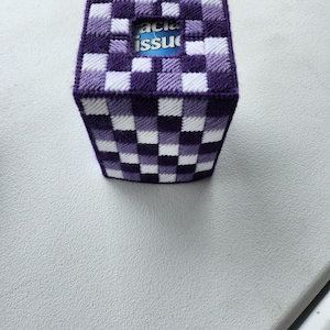 May include: A handmade tissue box cover in a checkerboard pattern of white and shades of purple. The opening reveals the words "facial tissue". The box is sitting on a white surface.