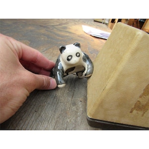 May include: A ceramic panda figurine with black and white markings. The panda is in a crouching position, with its front paws extended. The figurine is on a wooden surface.