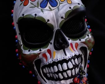 May include: A white skull mask featuring hand-painted floral motifs in red, blue, and yellow. The mask includes black mesh eye coverings, a black nose cavity, and a grinning jawline. The outer edges are decorated with small red and blue beads.