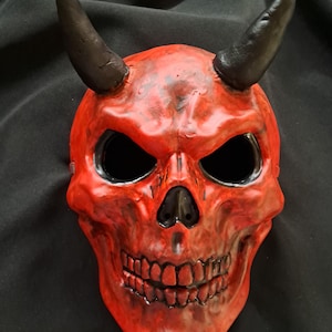 May include: A red skull mask featuring two prominent black horns extending from the top. The mask has deep, hollow eye sockets and a nasal cavity painted in black, with detailed teeth and a weathered, textured finish across the entire surface.