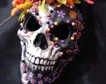 May include: A decorative skull mask, half silver and half black, adorned with colorful accents. The black side features purple flowers and orange and purple beads. The top is decorated with flowers and orange feathers.