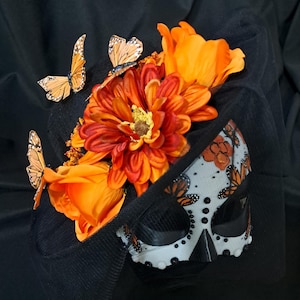 May include: A black hat adorned with vibrant orange artificial flowers and three monarch butterfly cutouts. A white mask with black detailing, butterfly motifs, and floral patterns is attached to the front, partially covered by black netting.