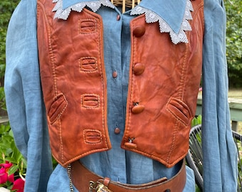 May include: A vintage ensemble featuring a rust-colored leather vest with button closures and a light blue collared shirt. The vest has decorative stitching and a fitted silhouette. A brown leather belt with a decorative antler buckle and chain completes the look. The outfit is displayed on a mannequin.