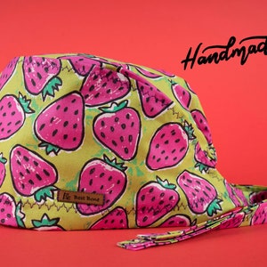 Op de afbeelding: Handgemaakte pet met een levendig aardbeienpatroon op een lichtgroene achtergrond. De aardbeien zijn roze met groene bladeren. De pet heeft een bijpassende band en een klein leren label met de tekst "Best Bone". Het woord "Handmade" is cursief geschreven.