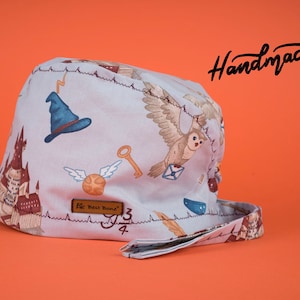 May include: A light blue surgical scrub hat with a whimsical Harry Potter themed print. The print features a Hogwarts castle, a golden snitch, a wizard's hat, an owl, and a key. The hat has a brown leather label that reads "For Best Bone".