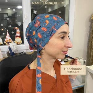 Op de afbeelding: Een op maat gemaakte scrubcap met een blauwe achtergrond en een patroon van oranje anatomische illustraties. De cap heeft een striksluiting en de tekst "CUSTOM SCRUB CAP" en "NAME EMBROIDERY". De tekst "Handmade with Care", "100% Cotton", "No Sweat" en "All Day Comfort" zijn ook zichtbaar.