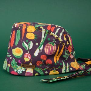 May include: A purple and green patterned surgical scrub hat with a long tie. The hat features a variety of vegetables, including carrots, onions, and peppers.