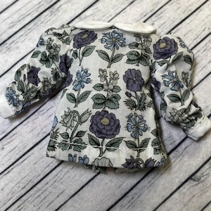 May include: A miniature floral print blouse with a white collar. The blouse features a pattern of blue and purple flowers with green leaves on a white background. The sleeves are long and cuffed. The blouse is designed for a doll.
