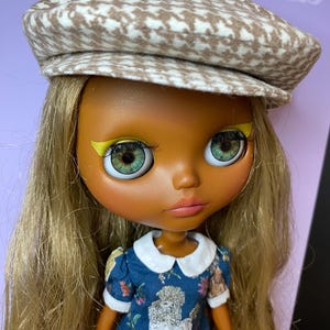 May include: A doll with long blonde hair and large green eyes wearing a brown and white houndstooth newsboy cap and a blue dress with a white collar. The dress features a poodle design.