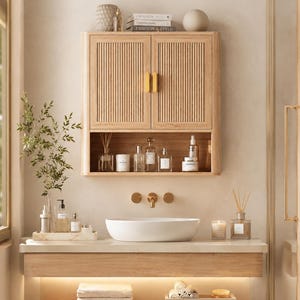 May include: A light wood wall-mounted bathroom cabinet with two vertical slatted doors and gold handles, featuring an open storage shelf below. The cabinet is positioned above a white oval vessel sink, a gold faucet, and a light wood vanity counter.