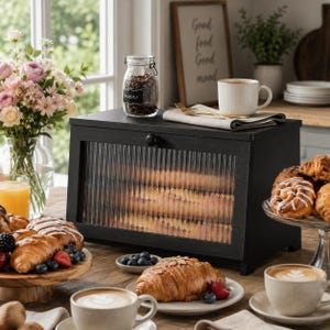 May include: A rectangular black bread box with a fluted glass front panel and a small round knob. The box sits on a wooden surface, topped with a glass jar labeled FUSILI, a white mug, and a folded napkin with a gold fork resting on top.