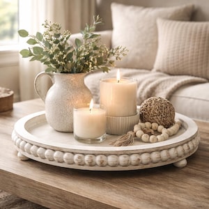 May include: A white, round, beaded tray holds a vase of greenery, two lit candles, a woven ball, and a strand of wooden beads. The tray sits on a wooden coffee table, creating a cozy, neutral-toned home decor scene.