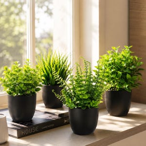 May include: Four artificial green plants in matte black rounded pots. The plants include two leafy shrubs, one tall grass-like plant, and one fern-like plant. They are arranged on a surface near a window, with one pot resting on a book titled HOUSE.