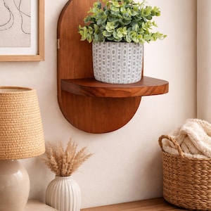 May include: A wooden wall shelf with a semi-circular design, holding a potted plant with green foliage. The shelf is mounted on a light-colored wall, with a framed artwork and a lamp visible in the background. A woven basket and a vase are also present.