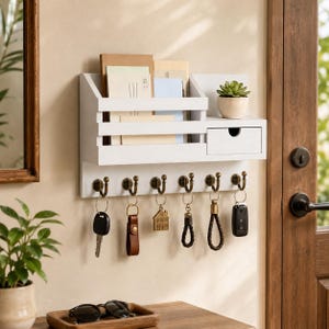 May include: A white wooden wall organizer with a shelf, drawer, and key hooks. It holds mail, a small potted plant, and keys with various keychains. The hooks are bronze-colored.