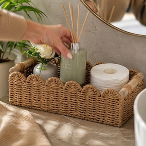 May include: A woven wicker tray with a scalloped edge holds a green glass diffuser bottle with reeds, a roll of white toilet paper, and a small vase with a white rose. The tray has wooden handles and is placed on a bathroom counter.