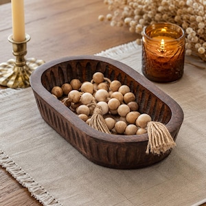 May include: A dark brown, wooden decorative bowl filled with light brown wooden beads and two beige tassels. A lit candle in a gold holder and a lit amber glass candle holder are in the background.