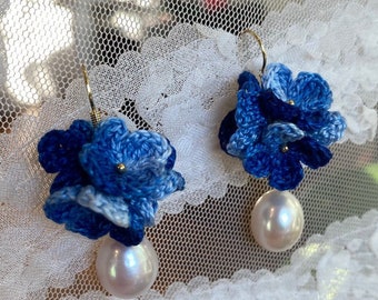 May include: A pair of blue crocheted flower earrings with a pearl dangle. The earrings are on a white lace background.