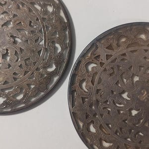 以下が含まれることがあります： 複雑な透かし彫りの花柄が施された、丸い銀色の金属製の壁飾り2つ。装飾品は円形で、渦巻く葉と花の精巧なデザインが特徴です。金属はわずかに古びた外観で、ヴィンテージ感を高めています。