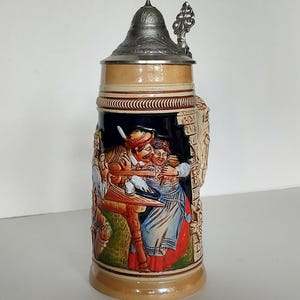 May include: A ceramic beer stein featuring a metal lid with a thumb lever. The body is tan with a central relief scene depicting two figures in colorful clothing sitting at a table. The background of the scene is black, and the side shows a textured stone wall design.