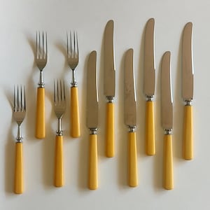 May include: A set of vintage silverware, including forks and knives. The utensils have stainless steel blades and tines, with bright yellow handles. The arrangement includes three forks and six knives.