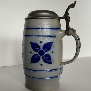 May include: A gray salt-glazed stoneware beer stein featuring a textured surface, a cobalt blue floral motif, and horizontal bands. The vessel includes a hinged pewter lid with an ornate thumb lift and a curved handle with blue accents.
