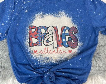 Peut inclure: Un t-shirt bleu avec un motif de blanchiment blanc du mot "BRAVES" avec un ballon de baseball et le mot "atlanta" en rouge, blanc et bleu.