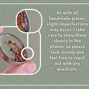 May include: Oval pendant with a clear resin dome, showcasing miniature mushrooms and earthy elements. The pendant has a silver-colored metal frame. The image includes text about handmade imperfections.