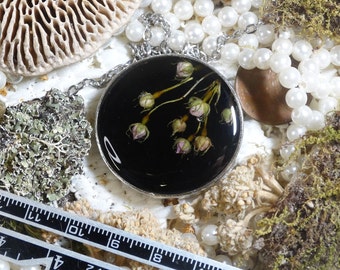 May include: A round pendant necklace with a black background, featuring small pink flower buds. The pendant is encased in a silver-colored metal frame and hangs on a silver chain. The background includes white pearls and natural elements.