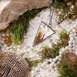 May include: A silver-toned pendant necklace with a clear, triangular resin pendant containing dried plants and a small mushroom. The necklace rests among white pearls, green moss, and a bronze unicorn figurine. The scene is set in a natural, earthy display.