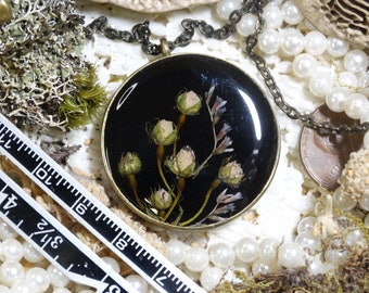 Puede incluir: Un collar con colgante redondo con fondo negro, con pequeñas flores claras preservadas. El colgante está encerrado en un marco de metal color bronce y cuelga de una cadena a juego. En el fondo se ven perlas y musgo.