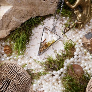 May include: A silver-toned necklace with a triangular pendant filled with dried plants and seeds. The pendant rests among pearls, green moss, and a small brass unicorn figurine. The background includes rocks and a textured surface.