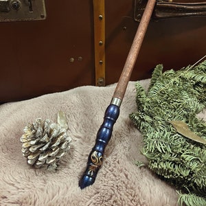 Pode incluir: Uma varinha de madeira marrom e azul escuro com detalhes dourados e um design decorativo. A varinha est&aacute; sobre uma superf&iacute;cie marrom e macia.