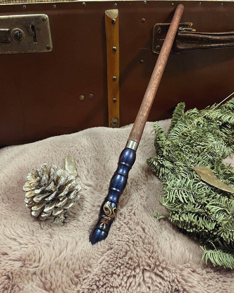 Pode incluir: Uma varinha de madeira marrom e azul escuro com detalhes dourados e um design decorativo. A varinha est&aacute; sobre uma superf&iacute;cie marrom e macia.