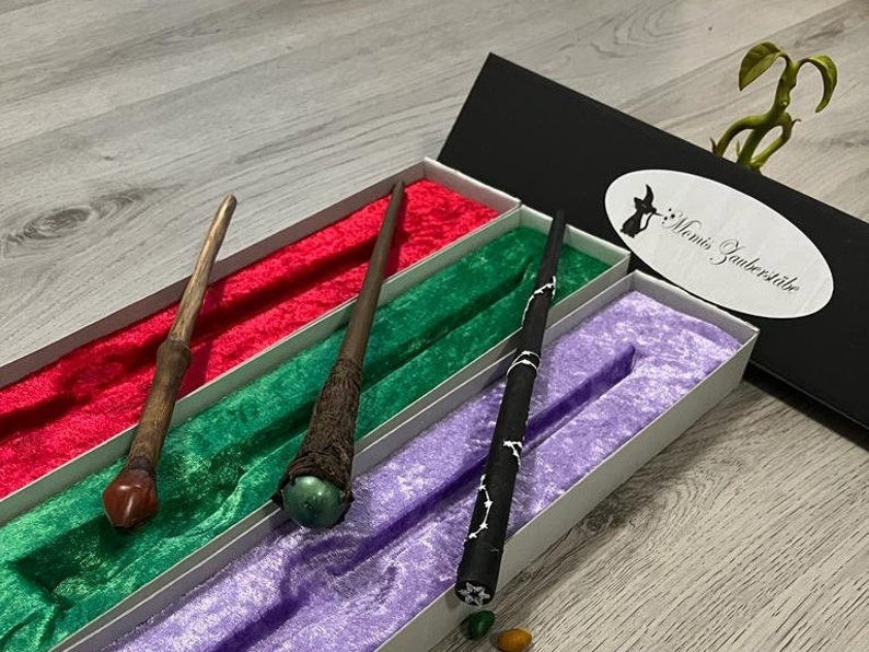 Pode incluir: Tr&ecirc;s varinhas de madeira com pontas de diferentes cores s&atilde;o exibidas em uma caixa forrada de veludo. A caixa &eacute; preta com um r&oacute;tulo branco que diz "Mami's Zauberstube".