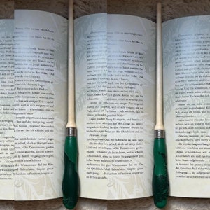 Pode incluir: Um close-up de um livro com um marcador de p&aacute;gina de madeira. O marcador &eacute; verde e branco com um design detalhado. O livro est&aacute; aberto em uma p&aacute;gina com texto em alem&atilde;o.