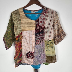 May include: A short-sleeve button-down blouse featuring a patchwork design of various floral, paisley, and geometric patterns in shades of green, red, cream, and purple. The garment has a round neckline and a bright blue interior lining.