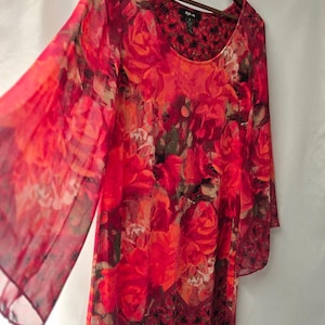 May include: A long-sleeved tunic featuring a vibrant red and orange floral print. The garment has a scoop neckline, sheer flowing sleeves, and a dark patterned border along the hemline. It is displayed on a wooden hanger against a white background.