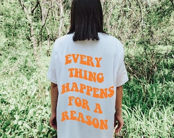 May include: White t-shirt with the orange text "EVERYTHING HAPPENS FOR A REASON" in a retro font. The shirt is worn by a person standing outdoors with a green, natural background.
