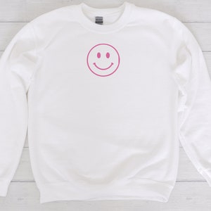 May include: White sweatshirt with a pink smiley face graphic on the front.